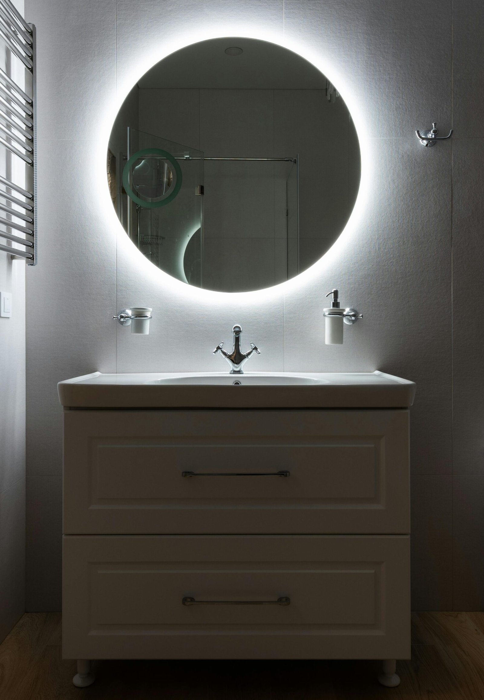 Modern bathroom with washbasin and dispenser bottle under round shaped mirror on light wall in house